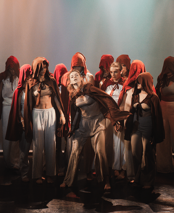 Groupe de danseurs en capes rouges sur scène, éclairage dramatique.