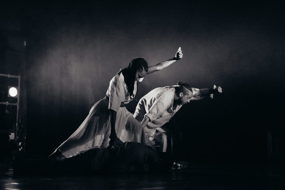 Danseurs en mouvement sur scène, éclairage dramatique en noir et blanc.