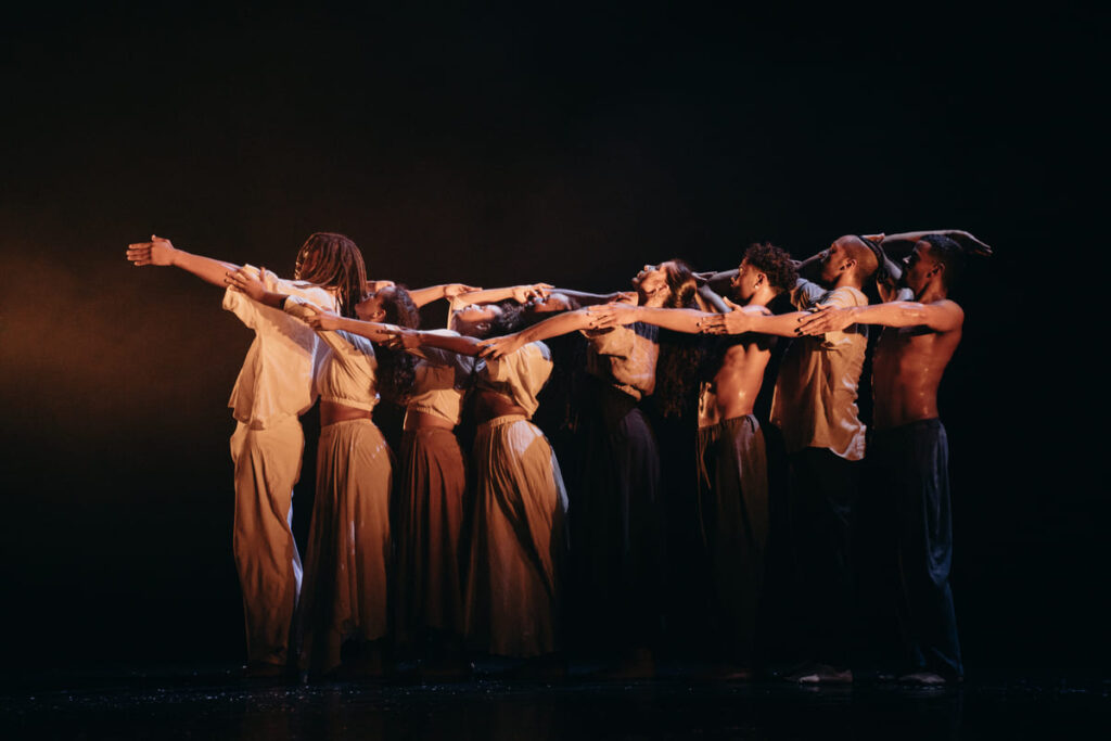 Groupe de danseurs en formation, bras tendus, éclairage dramatique.